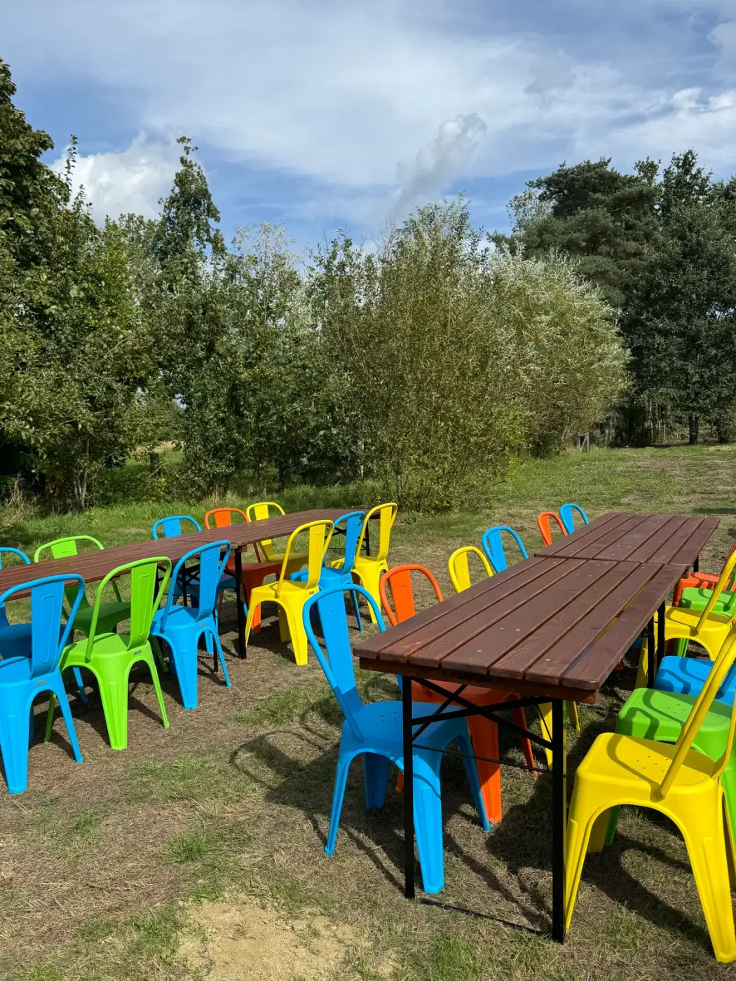 Lange feesttafels met kleurrijke stoelen op een grasveld onder een blauwe lucht, perfect om meubilair te huren voor een buitenfeest in Deurne
