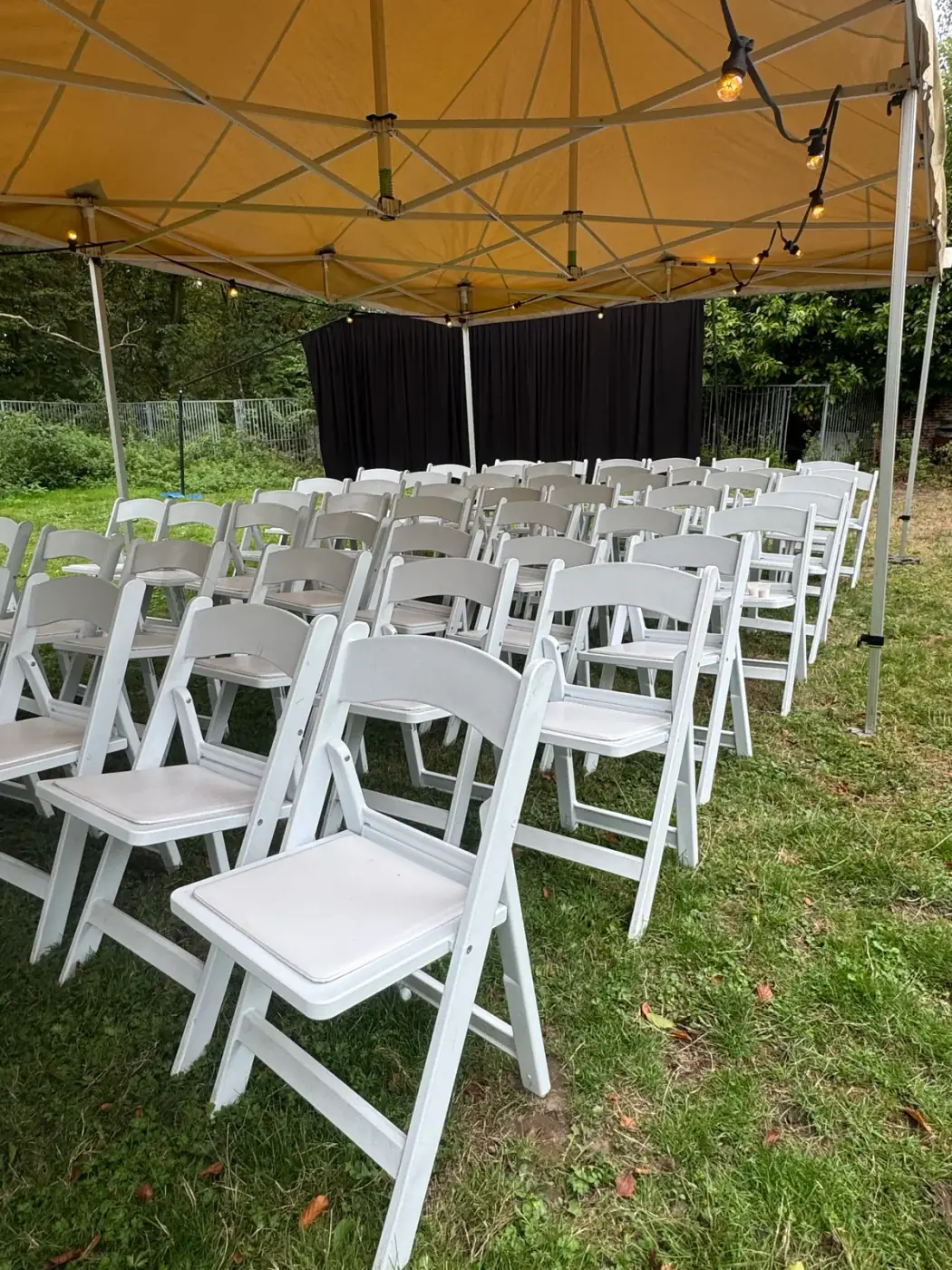 Witte klapstoelen onder een vouwtent met lichtslinger en een zwart achtergronddoek, sfeervol om meubilair te huren voor een ceremonie in Lier