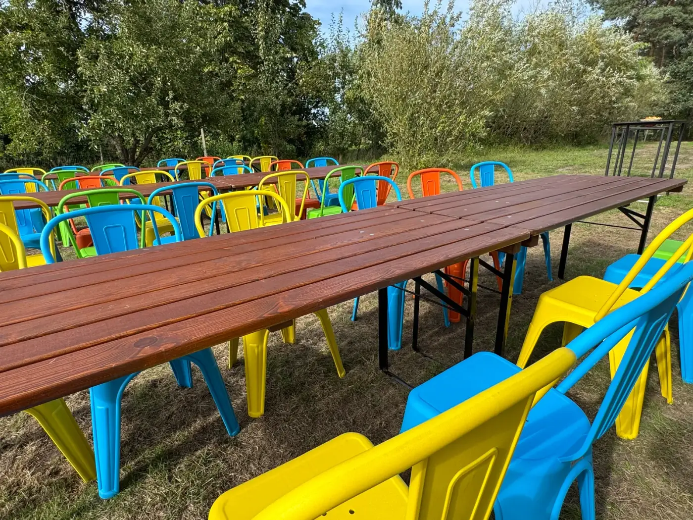 Twee lange houten tafels met blauwe, gele en groene stoelen klaargezet in de wei, ideaal om meubilair te huren voor een familiefeest in Mechelen