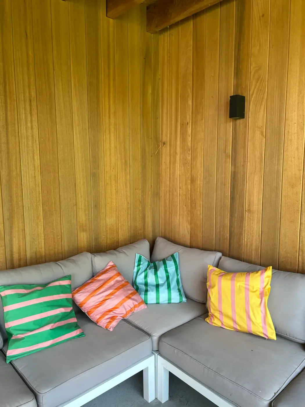 Grijsbeige hoekloungebank met gestreepte sierkussens tegen een houten wand in een overdekte buitenruimte, gezellig om meubilair te huren voor een loungehoek in Schilde