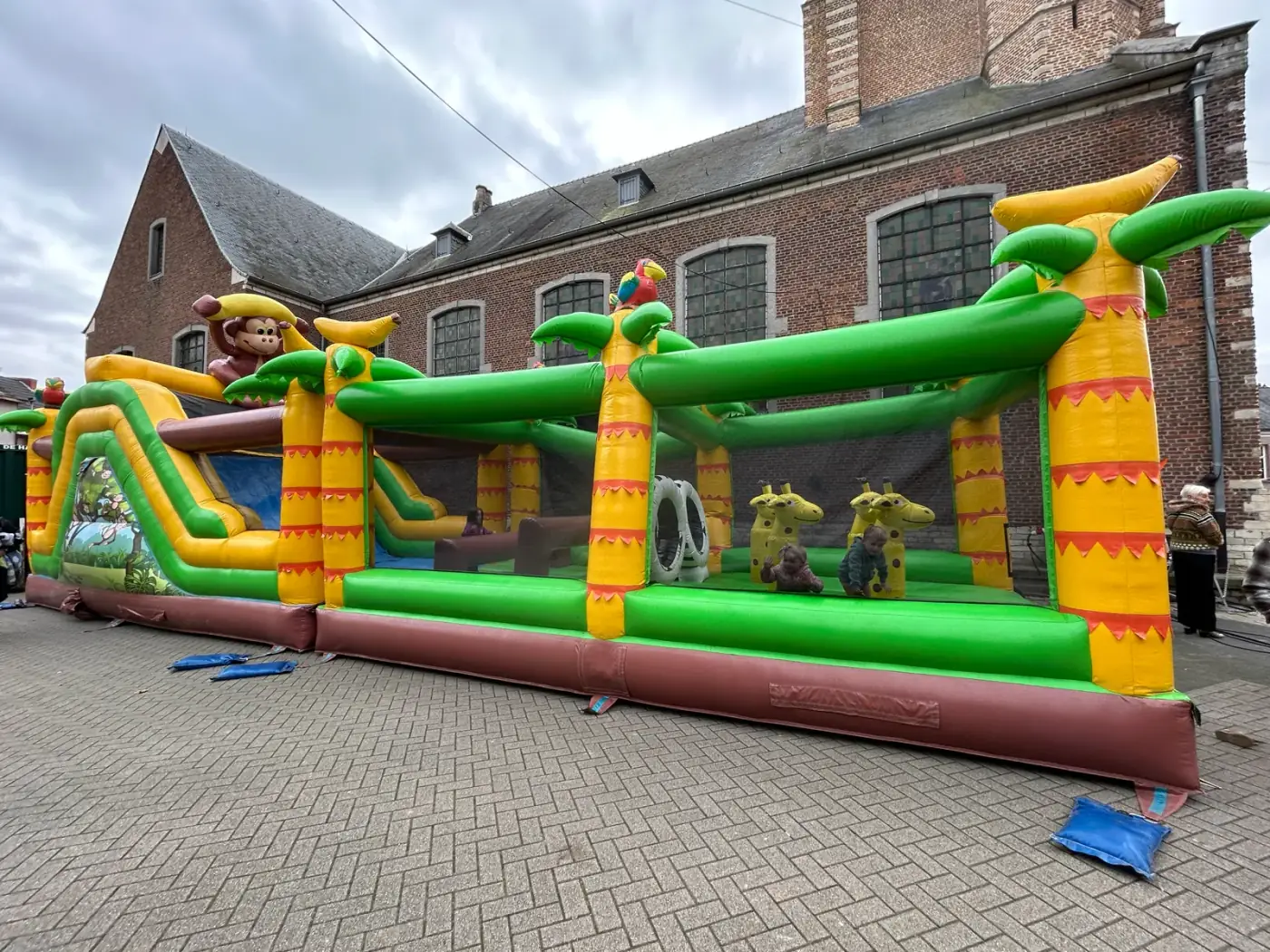 Groot springkasteel met junglethema en glijbaan op een geplaveid dorpsplein naast een kerk, klaar om een springkasteel te huren voor een dorpsfeest in Schilde