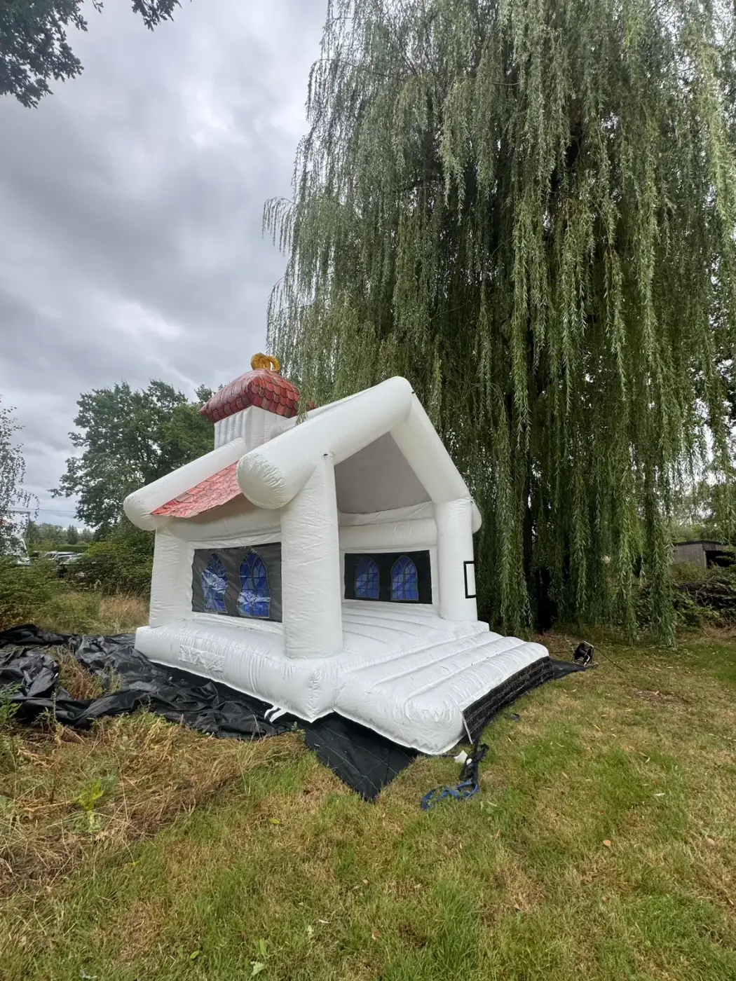 Wit springkasteel in de vorm van een huisje onder een treurwilg in de tuin, geschikt om een springkasteel te huren voor een communiefeest in Duffel