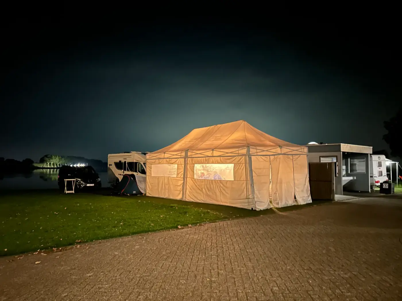 Verlichte beige vouwtent met raampanelen aan de waterkant bij valavond, sfeervol om een tent te huren voor een avondevenement in Ranst