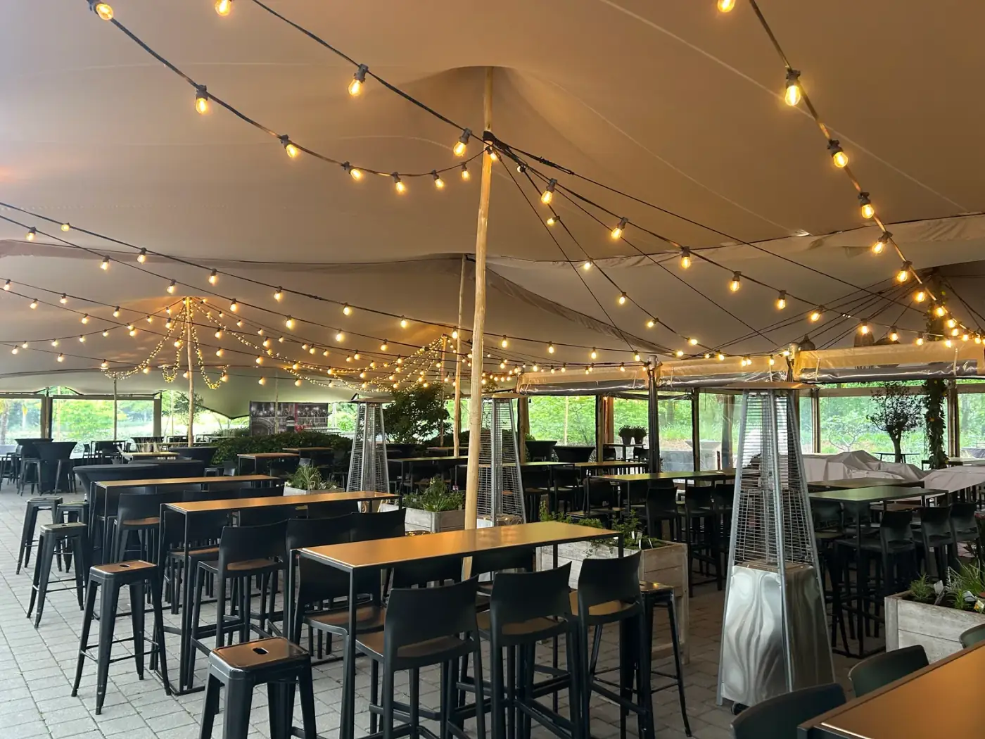 Piramidevormige gasterrasverwarmers tussen zwarte statafels onder een stretchtent met lichtslingers, onmisbaar om verwarming te huren voor een najaarsfeest in Ranst